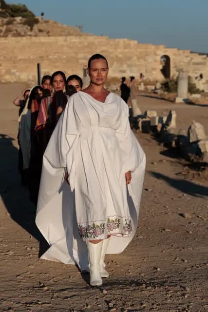 In a voluminous white dress with floral embroidery at the hem, a model opens the show along the earthen path of Knidos, the woven collection lined up behind her.
