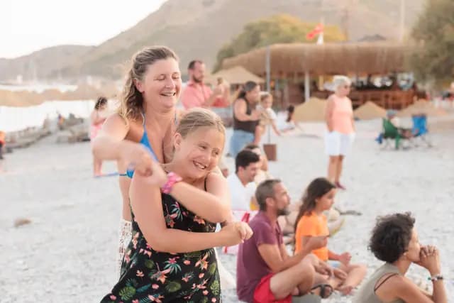 Two women dance and laugh in each other's arms on the pebble beach while the seated audience watches.