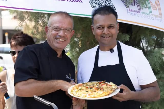 Two chefs hold up the finished plate together, smiling, the festival banner clearly readable behind them.