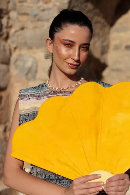 Holding a large yellow fan under her chin, a model gives the camera a warm, direct smile.