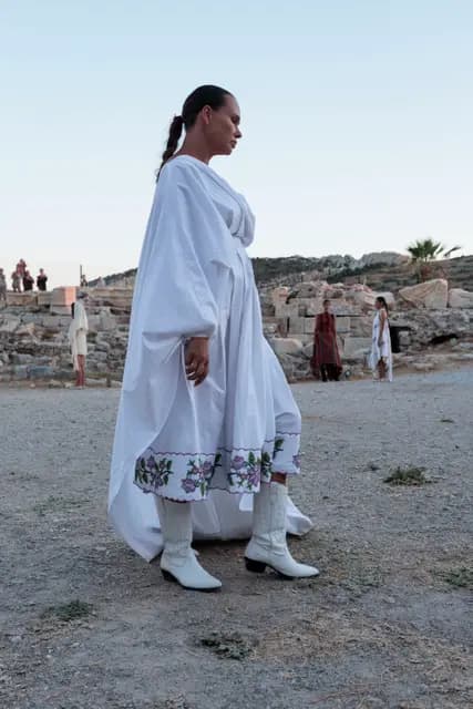 A model walks the earthen runway of Knidos in a long embroidered white dress.