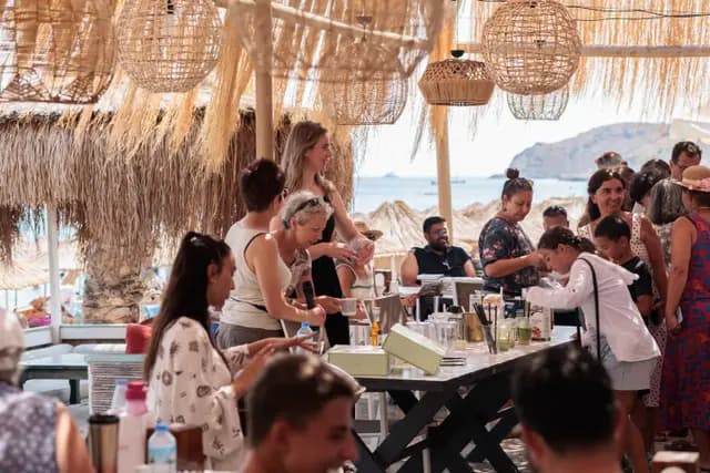 A crowd surrounds the workshop table under the reed canopy, the Datça bay and parasols behind.