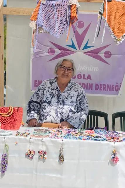 A veteran maker at the Muğla women's association stand looks straight into the lens beside her handmade jewellery.