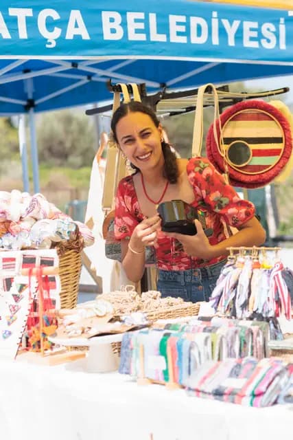 A young maker laughs warmly, glass in hand, at a stand full of handmade bags and weavings.