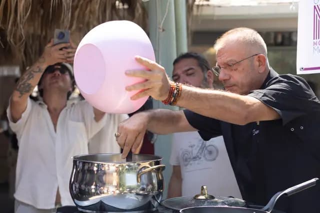 The chef empties a pink bowl into the large pot, the motion frozen at its peak.