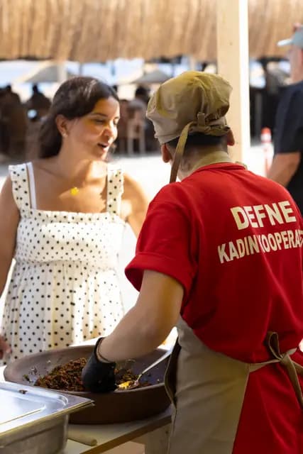 A cooperative member stirs a mixture in an earthenware pot while a visitor watches, smiling; the cooperative's name is readable on her back.