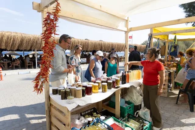Under strings of dried peppers, a producer explains her goods to visitors at a stand facing the sea.