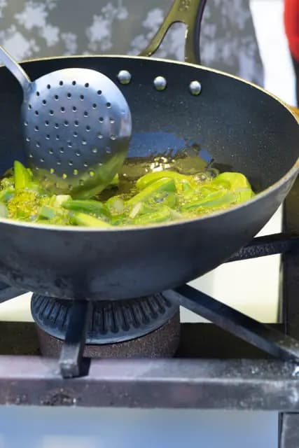 Green peppers frying in olive oil are turned with a skimmer in the iron pan.