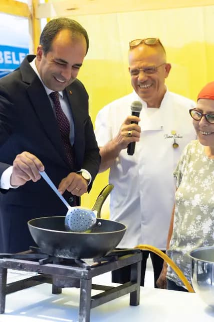 An official dips a ladle into the pan while the chef narrates on the microphone and the producer beside them laughs.