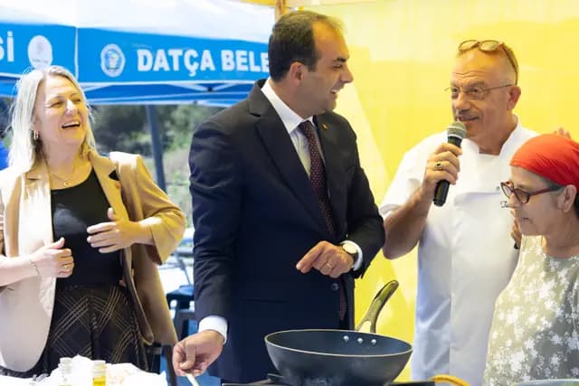 During the cooking show the chef narrates while officials and guests lean over the pan, laughing.