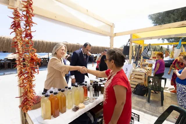 Under strings of red peppers, an official shakes hands with a woman producer at her stand, the Palamutbükü shore behind them.