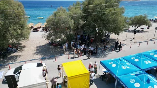 An aerial wide shot: the turquoise bay, the festival grounds under pine shade and the municipal parasols together.