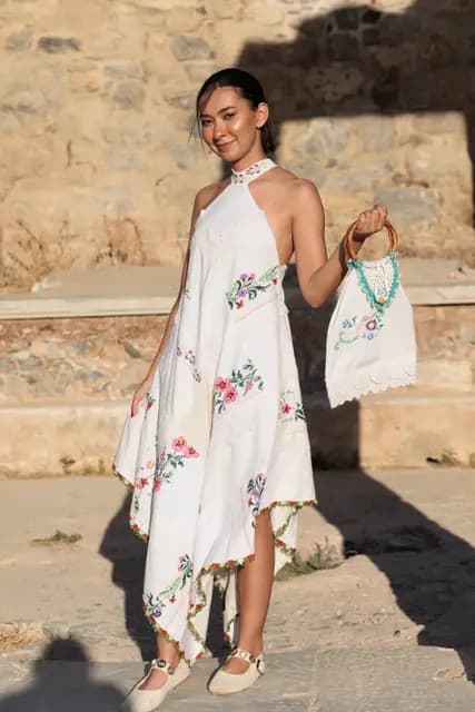 In a dress built from embroidered white linen panels, a model smiles at the camera showing off her embroidered bag.