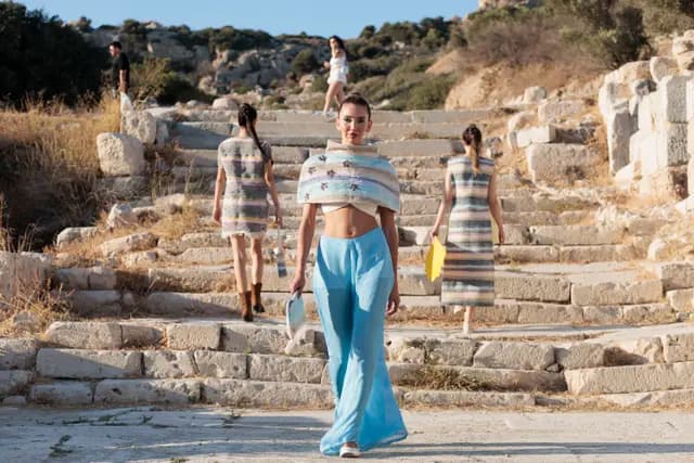 A model steps forward down the ancient stairs while the others carry on behind — the Knidos stage in full view.