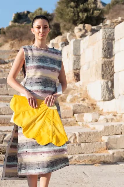 In a striped woven dress, a model walks towards the camera before an ancient wall, a yellow fan at her waist.