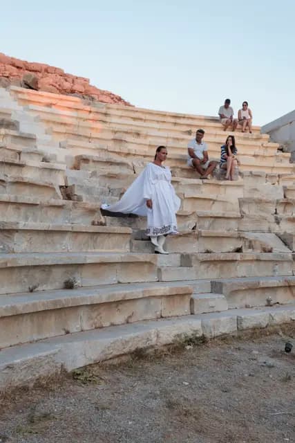 A model in an embroidered white dress descends the steps of the ancient theatre between the seated audience.