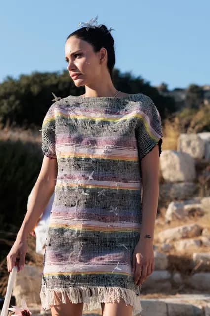 With a bird ornament in her hair, a model in a tasselled woven dress looks aside against the maquis.