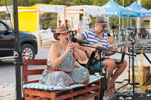 Two musicians on the shore: she plays a wind instrument while he accompanies on guitar.