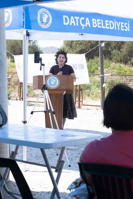 A speaker presents at the lectern, a phone tripod set up beside her to record.