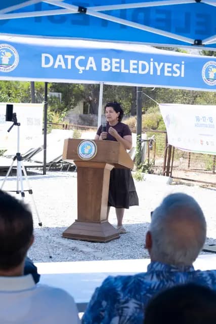 A speaker at the lectern, the festival poster and municipal canopy behind her.