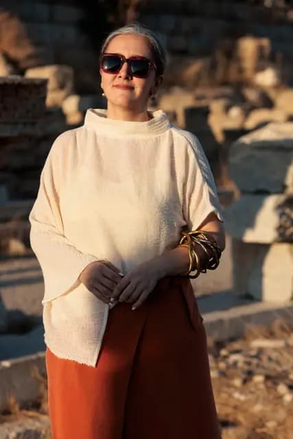 A model in a cream tunic and terracotta skirt, fragments of ancient columns behind her.