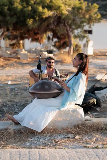 A musician sits among the stones of Knidos playing the handpan, the bay visible behind.